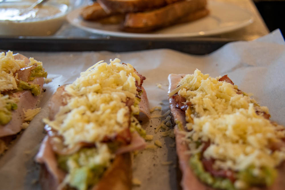 Toasted bread slices on a baking sheet with ham, bacon, avocado and mustard and cheese on top